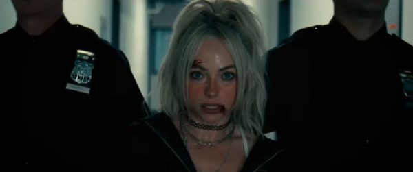 Liv Morgan appears disheveled with a cut on her forehead, flanked by two police officers as she is being arrested. The background features a dimly lit hallway, suggesting a tense situation.