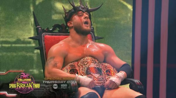 A muscular man wearing a crown sits on a throne, holding a championship belt in front of him. He appears to be celebrating victory after a wrestling match.