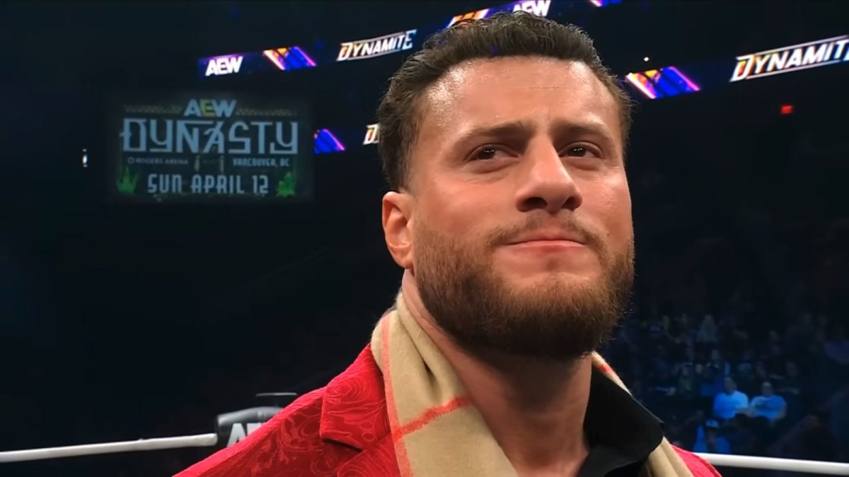 A wrestler with a serious expression is shown, wearing a red jacket and a scarf, during a tense moment in a wrestling ring. In the background, a promotional sign for AEW Dynamite and an upcoming event is visible.