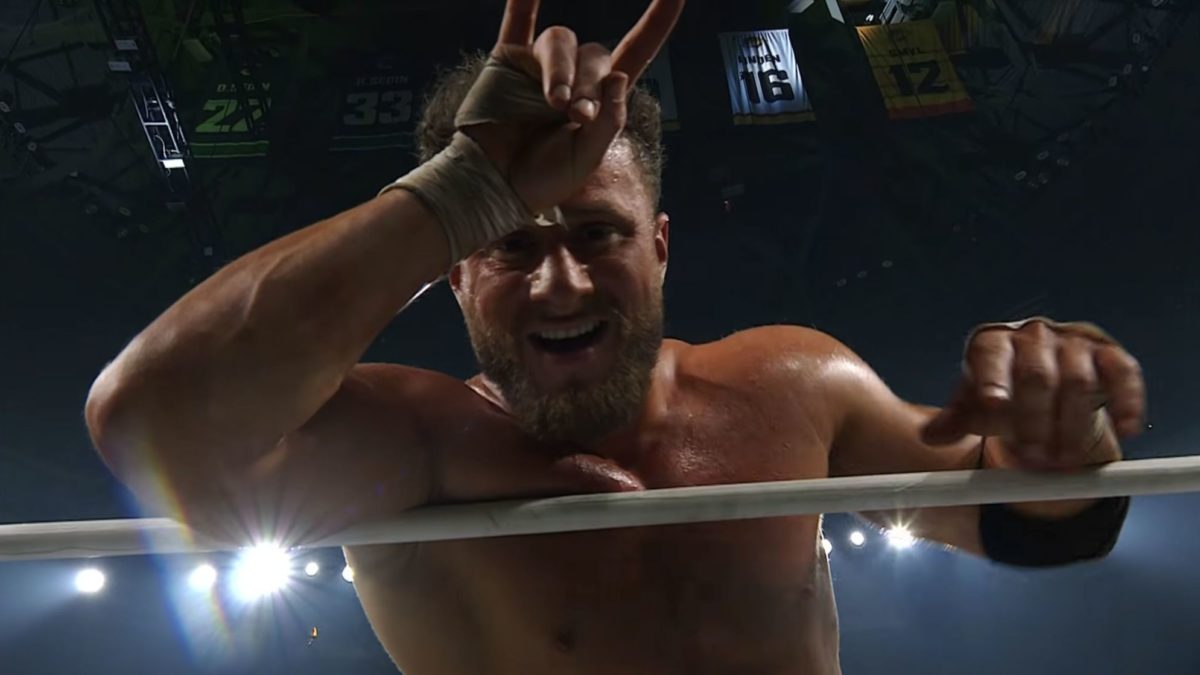 A wrestler smirks and holds up a hand gesture while standing in a wrestling ring, showcasing his confidence during a title match. The background is illuminated with bright lights and overhead banners.
