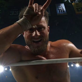 A wrestler smirks and holds up a hand gesture while standing in a wrestling ring, showcasing his confidence during a title match. The background is illuminated with bright lights and overhead banners.