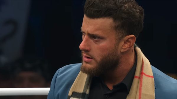 MJF, a professional wrestler, looks surprised and concerned while wearing a blue jacket and a scarf. His facial expression conveys intense emotion, likely reacting to an event during the AEW Dynamite Spring Breakthru event.