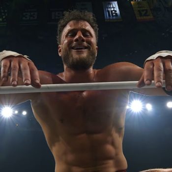 A wrestler with a beard smiles broadly while resting on the ropes of a wrestling ring, displaying a confident and villainous demeanor, with bright lights illuminating the scene above him.