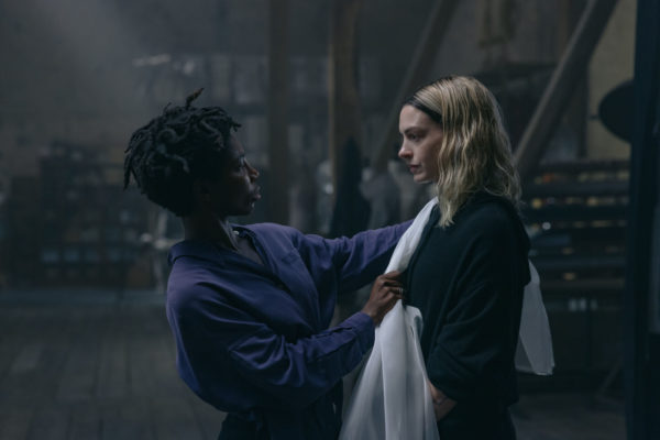 A dramatic scene featuring two women in a dimly lit room. One woman, wearing a purple shirt, is gently draping a white fabric around the shoulder of the other woman, who has long, wavy hair and is dressed in a dark garment.