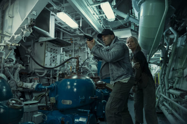 Jason Statham as Cole Reed and Annabelle Wallis as Angie Ellis are depicted in a tense moment within a dimly lit industrial setting, with complex machinery surrounding them; they appear ready to confront a threat.