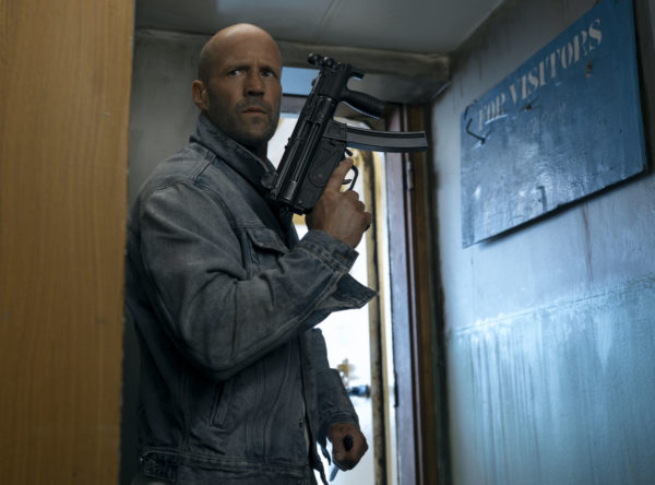 A tense moment from the movie 'Mutiny,' featuring a man in a denim jacket holding an assault rifle while peering around a doorway. A wooden sign on the wall reads 'FOR VISITORS,' adding to the scene's suspense.