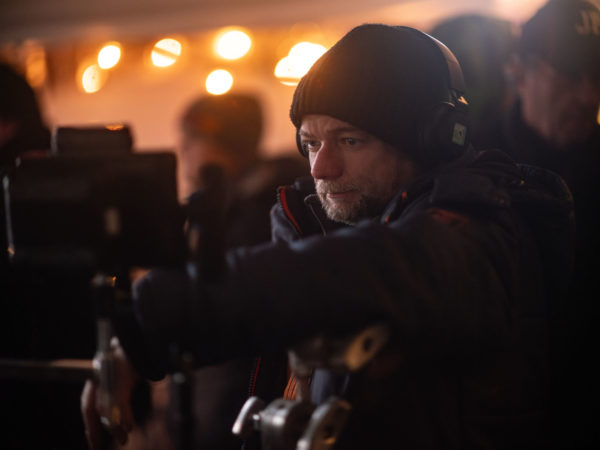 Director Andr&eacute; &Oslash;vredal is on the set of the film Passenger, focusing intently on the camera equipment amidst warm, blurred lighting in the background.