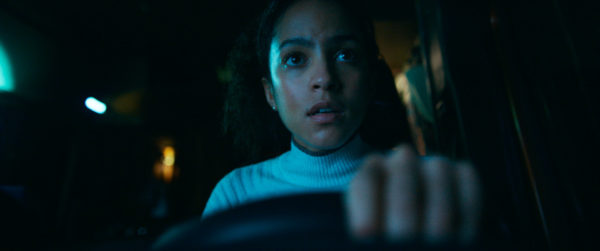 Lou Llobell portrays Maddie in a tense moment, gripping the steering wheel with a focused expression, set in a dimly lit vehicle interior.