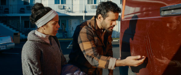 A scene from the film 'Passenger' featuring Lou Llobell as Maddie and Jacob Scipio as Tyler, standing in front of a red vehicle and appearing to examine a scratch on the surface. The background includes a building with visible outdoor steps and other parked cars.