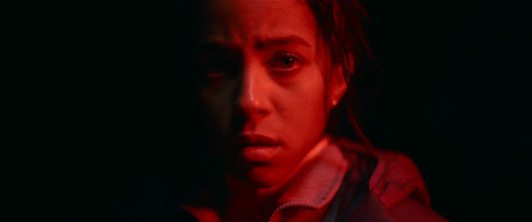 Close-up of Lou Llobell as Maddie in 'Passenger', featuring a dramatic expression illuminated by red lighting against a dark background.