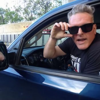 A man wearing sunglasses leans out of a vehicle window, seemingly in a playful or mischievous manner. He is dressed in a casual black t-shirt, and the background shows a parking area with trees.