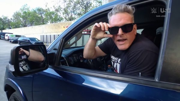 A man wearing sunglasses leans out of a vehicle window, seemingly in a playful or mischievous manner. He is dressed in a casual black t-shirt, and the background shows a parking area with trees.