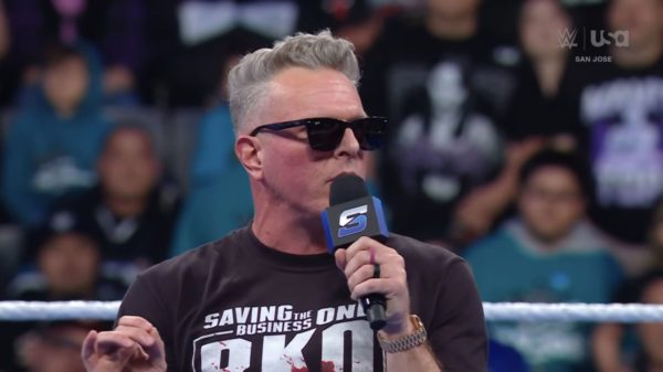 A man with gray hair and sunglasses is speaking into a microphone during a WWE SmackDown event. He is wearing a black t-shirt that says "Saving the Business" and looks engaged with the audience.