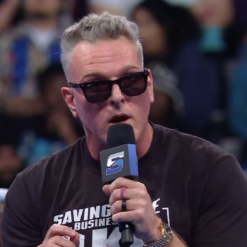 Pat McAfee speaks into a microphone while wearing sunglasses, standing in a wrestling ring. The audience is visible behind him, reacting to his taunts directed at the WWE Universe.