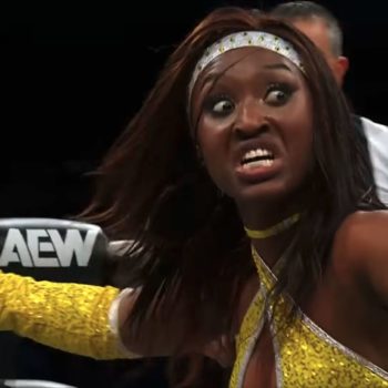 A professional wrestler, dressed in a sparkling yellow costume, passionately gestures and displays a fierce expression in the AEW Dynamite wrestling ring.