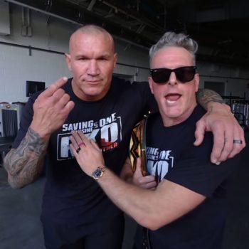 Randy Orton and Pat McAfee pose closely together in a backstage area, with Orton raising one finger, while McAfee has an excited expression and holds a WWE Championship belt. Both wear matching black T-shirts featuring the phrase 'Saving is One' and 'RKO'.
