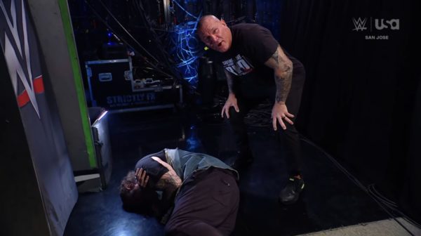 Randy Orton crouches next to a downed Cody Rhodes on the floor, surrounded by backstage equipment and dim lighting. Orton has an intense expression, clearly indicating a moment of confrontation after a move called the Jelly Roll.
