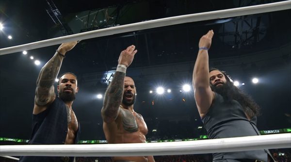 Three wrestlers celebrate their victory in the ring at AEW Dynasty, with intense expressions and raised arms. The atmosphere is energetic, with bright lights illuminating the scene.