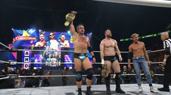 Three wrestlers celebrating in a wrestling ring after winning the AEW Trios Championship, with two holding championship belts. A colorful backdrop displays the event branding.