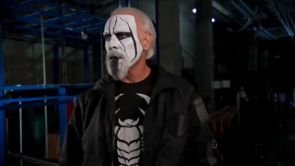 Sting, a professional wrestler, appears backstage with painted white face and dark markings, wearing a black jacket and a graphic t-shirt, as he prepares for an event.