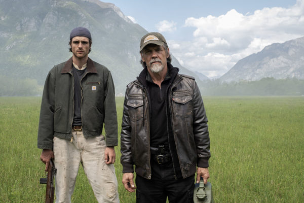 Two male characters, Jacob Elordi as Hig and Josh Brolin as Bangley, stand in a grassy field surrounded by mountains, reflecting a post-apocalyptic theme. Elordi holds a rifle, while Brolin carries a canister, hinting at their roles in the film "The Dog Stars."
