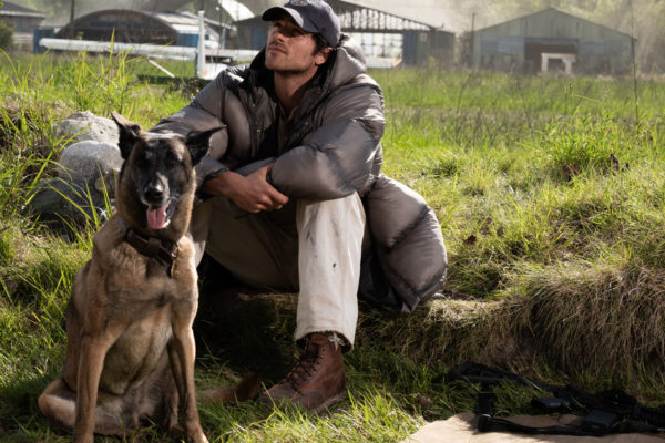A scene from 'The Dog Stars' featuring a man, dressed in a gray puffer jacket and rugged boots, sitting on the grass beside a dog. The dog has its tongue out and both appear to be gazing into the distance, surrounded by a rural backdrop.