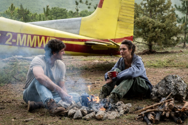 A scene from 'The Dog Stars' featuring Jacob Elordi as Hig and Margaret Qualley as Cima sitting by a campfire amidst nature, with a yellow and red airplane in the background. The characters are engaged in conversation, highlighting a moment of connection in a wild setting.
