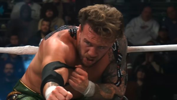 A professional wrestler, Will Ospreay, is in a dynamic pose, preparing to execute his signature move, the Hidden Blade, during a match at AEW Dynamite. He shows intense focus as he crouches against the ring ropes, wearing green and black wrestling gear, with fans visible in the blurred background.
