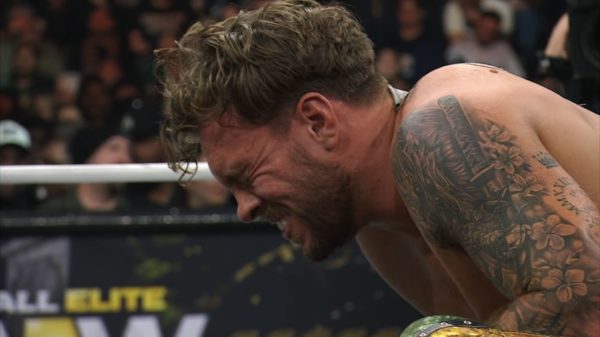 Will Ospreay, with long hair and tattoos, is shown grimacing in pain during his match against Jon Moxley at AEW Dynasty. The audience is visible in the background, capturing the intensity of the moment.
