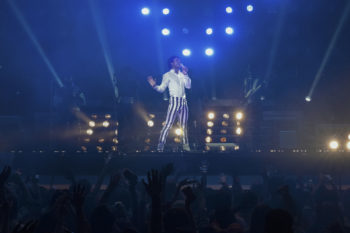 Jaafar Jackson performs as Michael Jackson on stage, wearing a white top and striped pants, with dramatic lighting and a cheering crowd in the background. The scene captures the energy of the biopic 'Michael'.