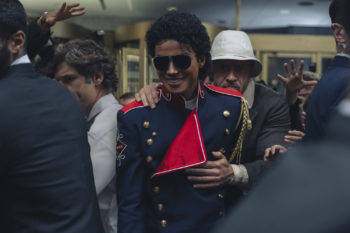 Jaafar Jackson portrays Michael Jackson in a blue military-style jacket with a red accent, surrounded by a crowd. The scene captures the intensity of a moment with hands reaching out, conveying a sense of excitement.