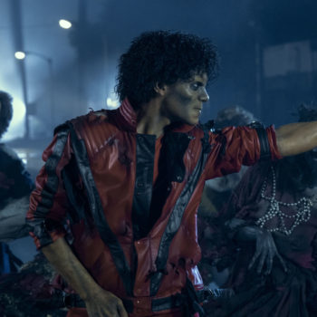 A still from the biopic 'Michael' featuring Jaafar Jackson portraying Michael Jackson, dressed in a tattered red jacket, performing amidst a group of zombie-like characters. The scene is set in a gloomy atmosphere with muted lighting.