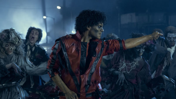 A still from the biopic 'Michael' featuring Jaafar Jackson portraying Michael Jackson, dressed in a tattered red jacket, performing amidst a group of zombie-like characters. The scene is set in a gloomy atmosphere with muted lighting.