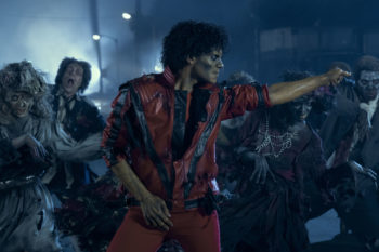 Jaafar Jackson portrays Michael Jackson in a dynamic scene, wearing a distinctive red jacket with black details, surrounded by a group of zombie-like figures. The atmosphere is moody and dark, reflecting a dramatic moment from the upcoming biopic 'Michael'.