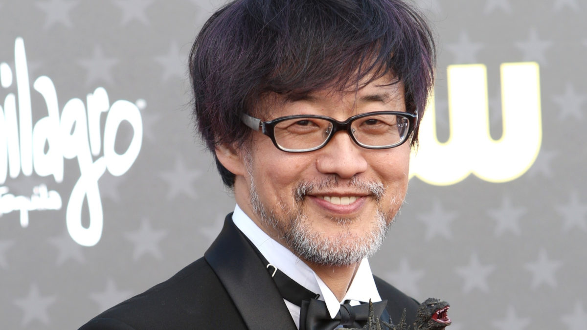 A man with glasses and facial hair smiles, holding a small Godzilla figurine, dressed in a formal black outfit with a bow tie. He stands against a backdrop featuring stars, promoting the movie 'Godzilla Minus One' directed by Takashi Yamazaki.