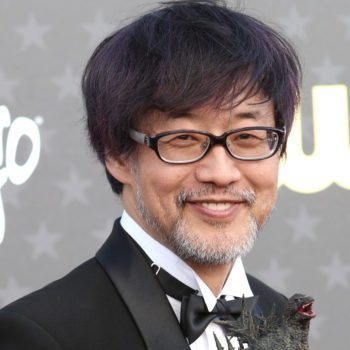 A man with glasses and facial hair smiles, holding a small Godzilla figurine, dressed in a formal black outfit with a bow tie. He stands against a backdrop featuring stars, promoting the movie 'Godzilla Minus One' directed by Takashi Yamazaki.