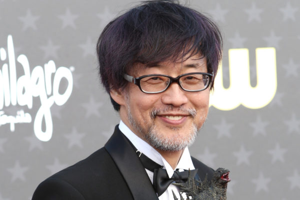 A man with glasses and facial hair smiles, holding a small Godzilla figurine, dressed in a formal black outfit with a bow tie. He stands against a backdrop featuring stars, promoting the movie 'Godzilla Minus One' directed by Takashi Yamazaki.