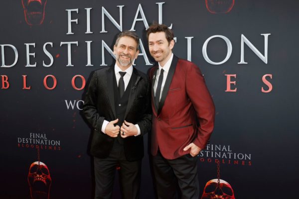 Two men, Adam Stein and Zach Lipovsky, pose together on the red carpet, wearing stylish suits, in front of a dark backdrop featuring the text 'FINAL DESTINATION BLOODLINES' and a graphic of a skull. The setting appears to be the premiere event for the movie.