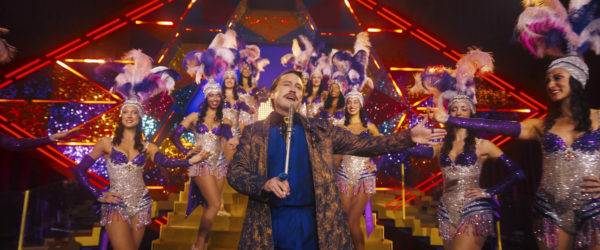Kieran Culkin as Caesar Flickerman performs on stage surrounded by dancers in glittering costumes, with vibrant lighting and a colorful backdrop in the scene from The Hunger Games: Sunrise on the Reaping.