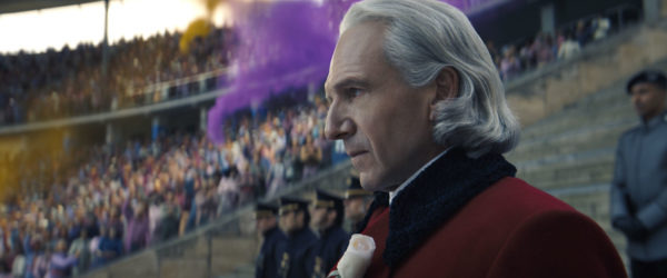A close-up of President Snow, portrayed by Ralph Fiennes, standing in a stadium filled with an audience, amidst colorful smoke in the background. He is dressed in a red coat with a white rose pinned to his lapel.