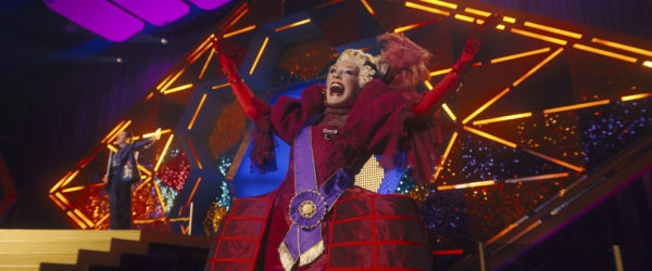 Drusilla Sickle, portrayed by Glenn Close, dramatically presents on stage with colorful, flashy lighting in the background, dressed in an elaborate red costume showcasing a mix of textures and bold accessories.