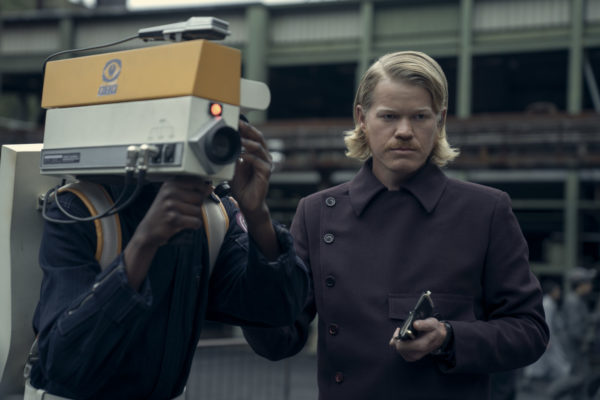 In a scene from The Hunger Games: Sunrise on the Reaping, a character portrayed by Jesse Plemons stands beside a person holding an old-style video camera. The setting appears industrial, with muted colors and a somber atmosphere.