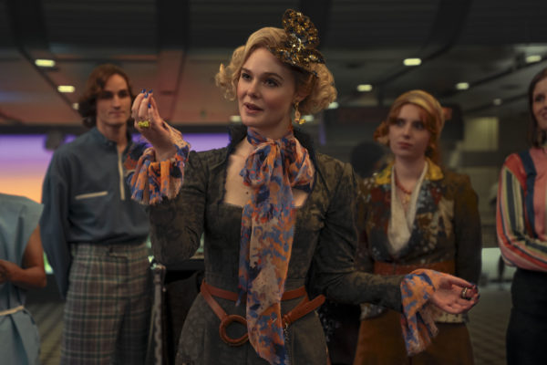 Elle Fanning portrays Effie Trinket in a colorful costume, complete with a floral scarf and decorative hairpiece, in a scene from 'The Hunger Games: Sunrise on the Reaping.' The background features additional characters in similarly vibrant attire.